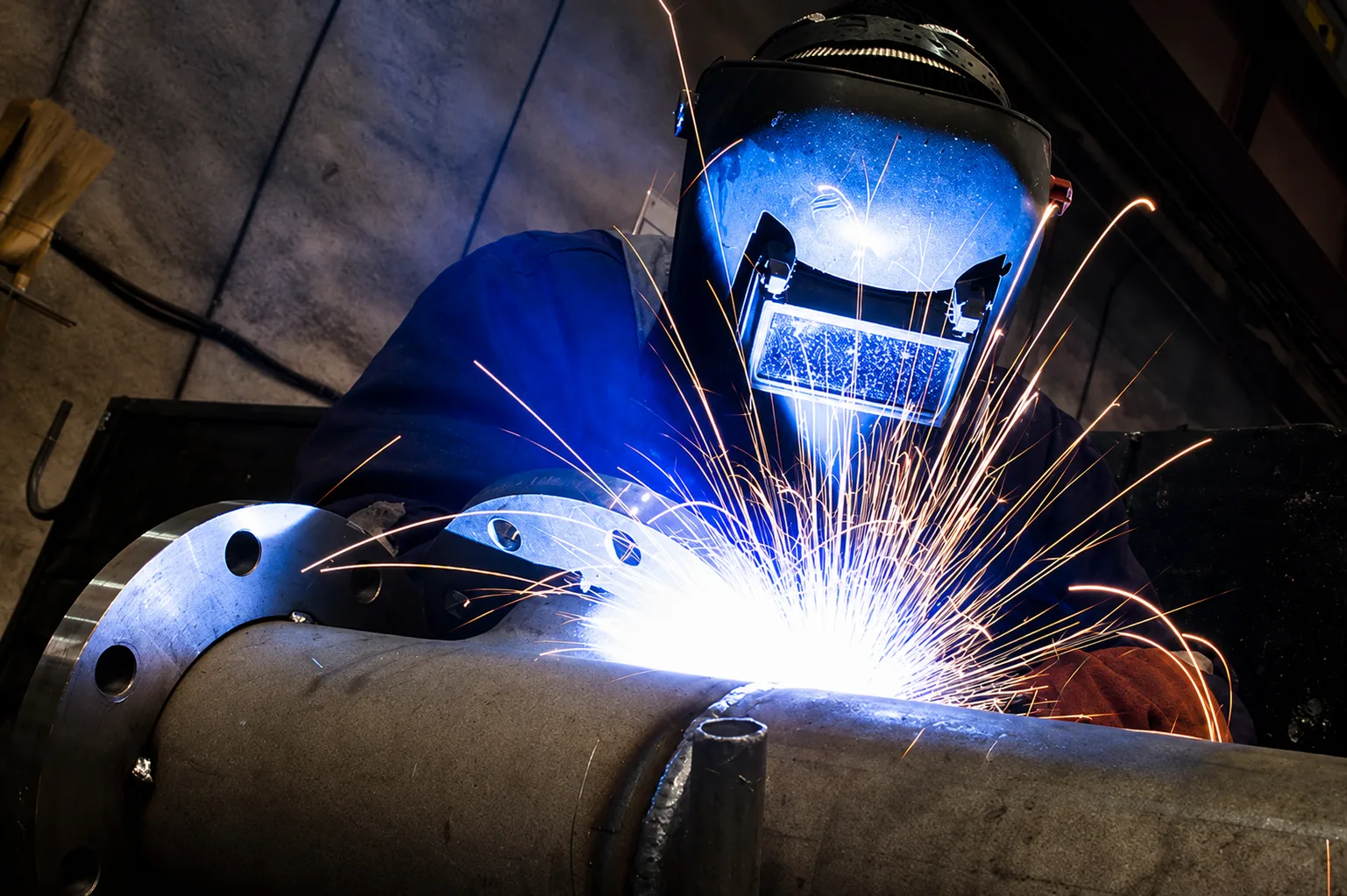 Welder producing sparks while welding a flanged industrial assembly